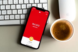 An iphone screen displays 164,913 YouTube subscribers and sits on a work desk together with an apple keyboard, mouse, and a cup of coffee.