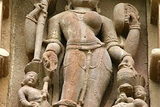 An image of a sculpture found in Khajurao where one side if female and the other male. There are four arms which carry various symbols of blessing and weapons. There are also two other short forms near the feet of the sculpture. the scultpure is made from stone and appears to be part of wall panel.