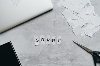 Black and white photo of a desk. The letters “SORRY” have been individually cut out and placed in the middle. There is a pair of scissors, paper scraps, a laptop, and a book around the letters.