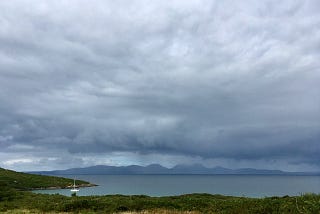 Low, purple clouds above the Paps of Jura — mountains on the Scottish island of Jura. There is a yacht anchored in blue/green calm sea.