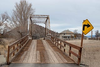 An old one-lane wood-and-iron bridge across a small river on a rural two-lane dirt road, with a farmhouse in the background.