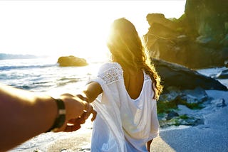 back view of woman with long brown hair wearing white dress walking on rocky beach holding man’s hand with arm outstretched behind her