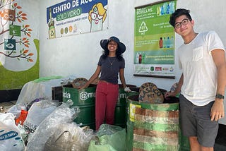 Two young adults pose in front of recycling waste bins.