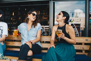 Two Smiling Women Sitting on Wooden Bench Drinking a Beer