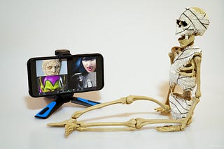 A Halloween skeleton sits in front of a computer screen that has a Zoom conference call showing four other Halloween characters that are very scary. The skeleton’s mouth is dropped open like he is in shock.