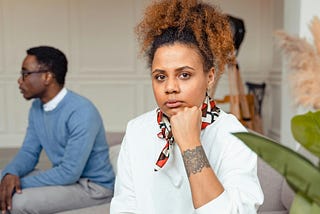 woman rest her chin on closed fist, man sits in background looking away