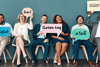 several diverse adults, sitting in a room. Each is holding up a sign with varying language greetings (Good day or Hello in French, Italian, German, Thai, Arabic)