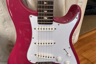 PRS Silver Sky SE guitar, in Dragonfruit color with white pickguard. Guitar is on guitar stand.