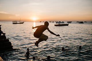 Image of a boy jumping off a dock into a lake at sunset. Other boys are in the water, playing. And there are two canoes, with paddlers, off in the distance.