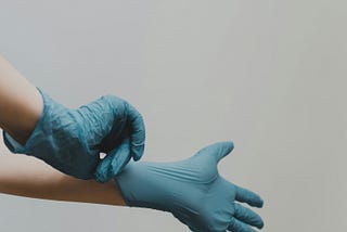 2 hands stretching a blue plastic doctor’s glove over the other hand.
