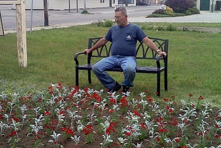 Grandfather sitting on wrought iron bench in a community garden of red and white flowers he planted, wearing blue jeans and navy t-shirt