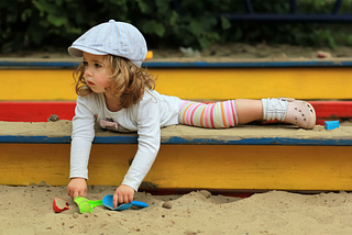 A young girl playing in a colorful wooden sandbox with colorful shovels.