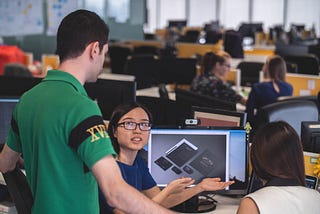 An office set up where a young man is looking over a colleague’s desktop while she is explaining her work to him.