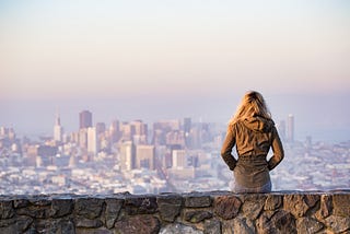 Woman sitting on a wall facing a city in the distance.