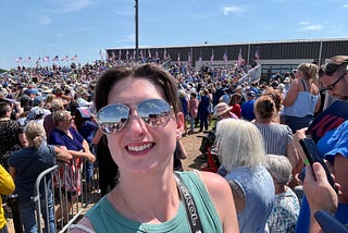 Allison stands wearing aviator sunglasses in front of a crowd of people on a sunny day. There are American flags in the distance.