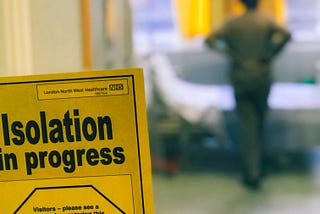 Quarantined room door with visuals of a patient standing inside. On the door a paper reading “isolation in progress”