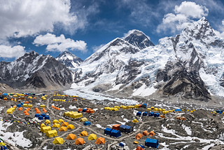 A mountaineering base camp on rocky glacial ground, with small tents dwarfed by a vast snow-covered peak rising into the sky above them.
