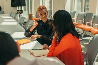 A Black Woman with strawberry blonde hair in sitting at a board room table signs to a South Asian woman with black hair wearing orange