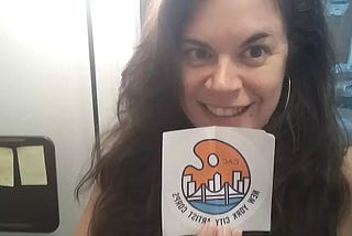 Author holds up New York City Artist Corps sticker with logo.