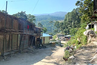 Rustic buildings align a dirt road that extends forward into the hilly countryside. Blue sky rises above dark green forested hills in the background.