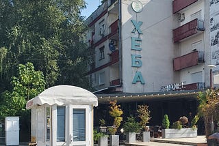 A street scens showing a tall apartment building with a sign in Cyrillic writing. In front in a small white kiosk. Trees surround all of it.