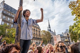 Woman standing on light post in the middle of protest and raising fist
