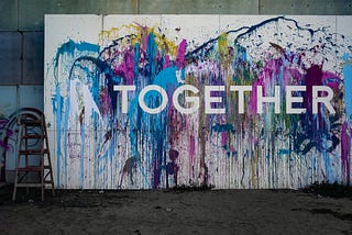 Pale blue, deep indigo, yellow and magenta paint are splashed onto a large white board, with the word ‘TOGETHER’ on it.