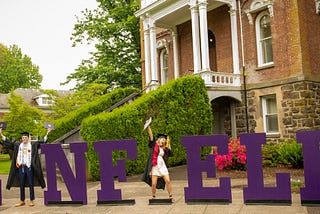 A man and woman in caps and gowns celebrating in front of a historic brick building.