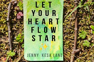 A watercolor rectangle I painted splashed with bright greens and yellows, on the green grass and moss, with the words, “Let Your Heart Flow Star” and my name Jenny Kesa Lane. There are four small sticks surrounding it like a border