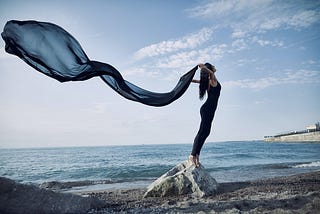 an image of a lady standing on a rock by the sea looking free and tranquil.