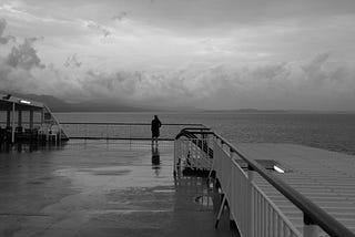 Black-and-white photo of a woman alone on a boat, heading off into the distance.