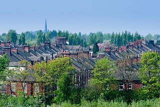 Terraces of houses near the city centre of Stoke on Trent, between lines of green-leaved trees. In the background, a church steeple rises above the treeline into a cloudless sky.