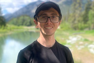 A close up photo of man smiling. Behind him, mountains and a river are visible.