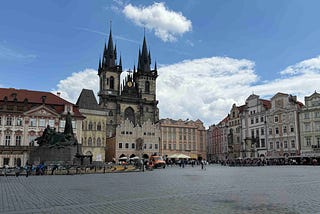A large historic square in Prague with cobblestone paving, people walking, and outdoor seating. The Gothic Church of Our Lady before Týn rises in the background with its two tall spires, and the Jan Hus Memorial stands prominently in the center, surrounded by ornate buildings with red‑tiled roofs under a partly cloudy sky.