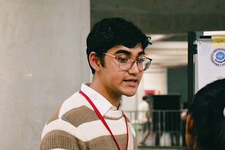 Vedant Dobwal speaking. He’s wearing a beige and tan striped sweater and a red lanyard.