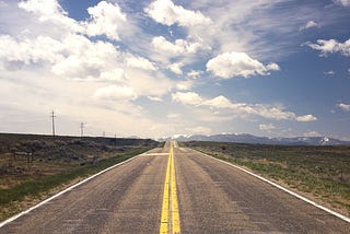Photo of a barren open road without any cars, just 2 lanes divided by yellow lines on the roadway with hydro poles and grass on the sides of the road and a blue sky with the sun shining down on the road.