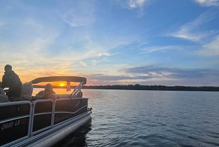 A pontoon is leaving the frame of the photo in the bottom left corner of the photo. You can see two people on the boat. A forested shoreline is in the distance and the sun is descending into the horizon behind the boat.