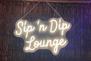 Photo of a neon sign, “Sip ‘n Dip Lounge” hanging on a chain on a rattan wall