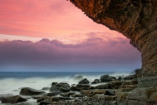 Edge of a sea cliff with rocks below, pink sky behind