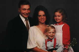 A family of four, left to right: a father wearing a tie, a mother wearing a white shirt with black hair, and a daughter with a red and white dress. In front of them is a little boy wearing a light blue shirt and red suspenders and a bow tie. In the lower right, there is a watermark noting that it was taken by Olan Mills in 1988.