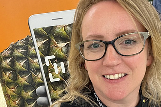 A photo of Vicky smiling in front of a photo of a pineapple being scanned by a phone.
