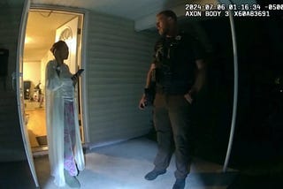 A nighttime body-camera image shows a woman standing in the doorway of a light-colored house, lit by warm indoor light. She wears a long robe, patterned pajama pants, and slippers, holding a phone to her ear. On the porch, a uniformed sheriff’s deputy in dark tactical gear faces her, partly in shadow, his flashlight beam cast on the floor.