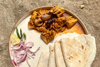In a plate, a flatbread along with meat curry is served with sliced onions, lemon, and green chillies on the side.