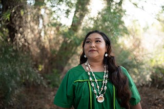 Andra Gutierrez standing with the green Gila River marshlands behind her. She is wearing a traditional O’otham attire with seashell jewelry.