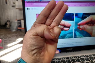 A close-up photo shows a person’s hand making a sign with their thumb and little finger touching. The other three fingers are extended straight up. In the background, there’s a blurred view of a laptop screen displaying what appears to be a webpage.