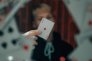 A blurred figure holding the ace of diamonds between their fingers at the viewer, surrounded by a whirling ring of playing cards