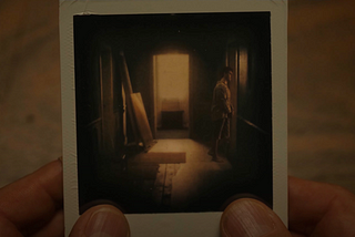 A close-up of a Polaroid photograph held in Philip’s hands, showing a young Philip standing alone in a dim hallway, bathed in warm light coming from an open door at the far end.