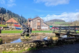 A winged metal dragon sculpture stands in a small pond beside a stone cottage and wooden footbridge, with green hills and buildings near Tintern Abbey under a bright blue sky.