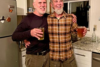 Two men stand in a kitchen with their arms around each other, holding up glasses of beer.