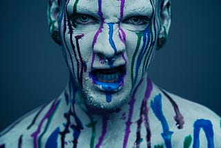 A growling man painted gray with drips of blue and red paint running down his face and out of his mouth